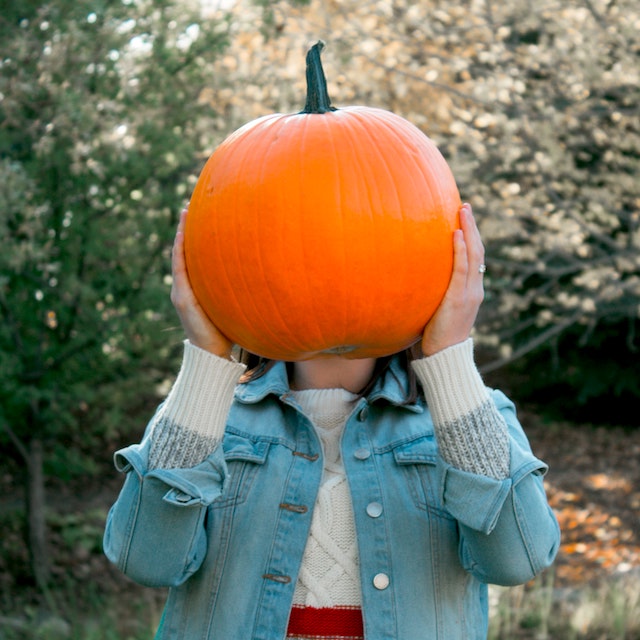 A picture of a girl wearing a sweater and jean jacket, holding an orange pumpkin over her face