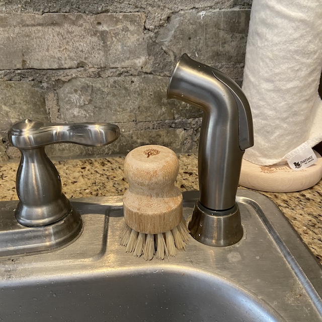 A picture of a bamboo dish brush sitting on a stainless steel kitchen sink