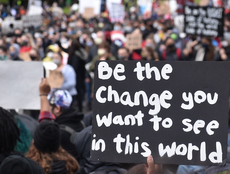 A picture of a protest with a hand holding a sign that says "Be the change you want to see in this world"