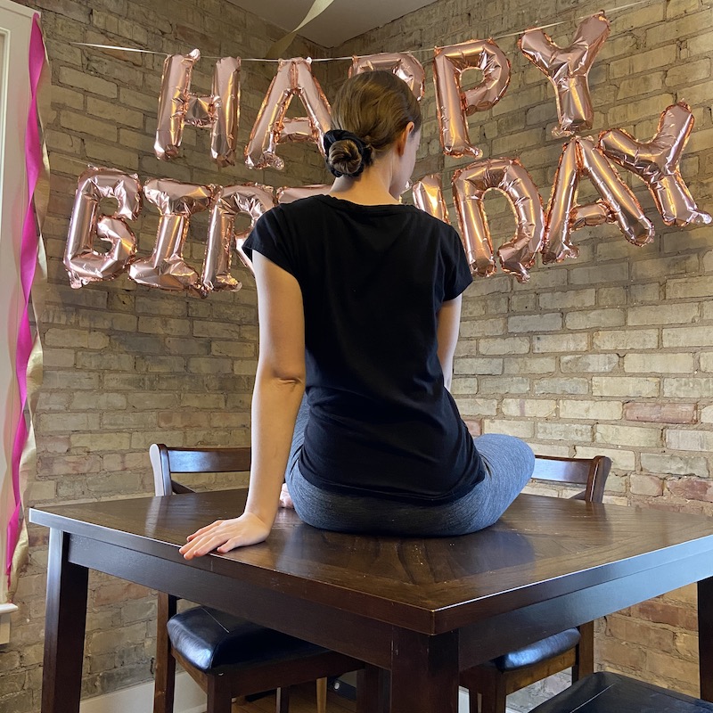 A picture of a girl sitting on a table in front of Happy Birthday balloons